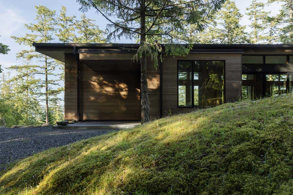 Low-profile cedar-clad home with flat roof overhang and garage entry on moss-covered hillside