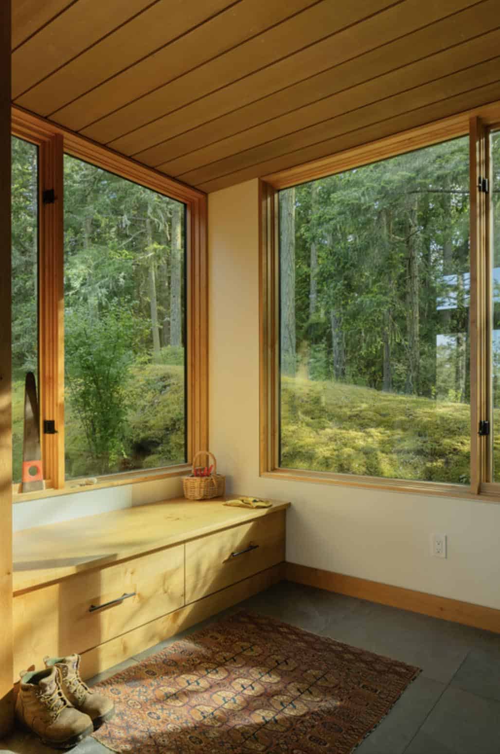 Built-in window seat with storage drawers beneath corner windows overlooking moss-covered forest floor