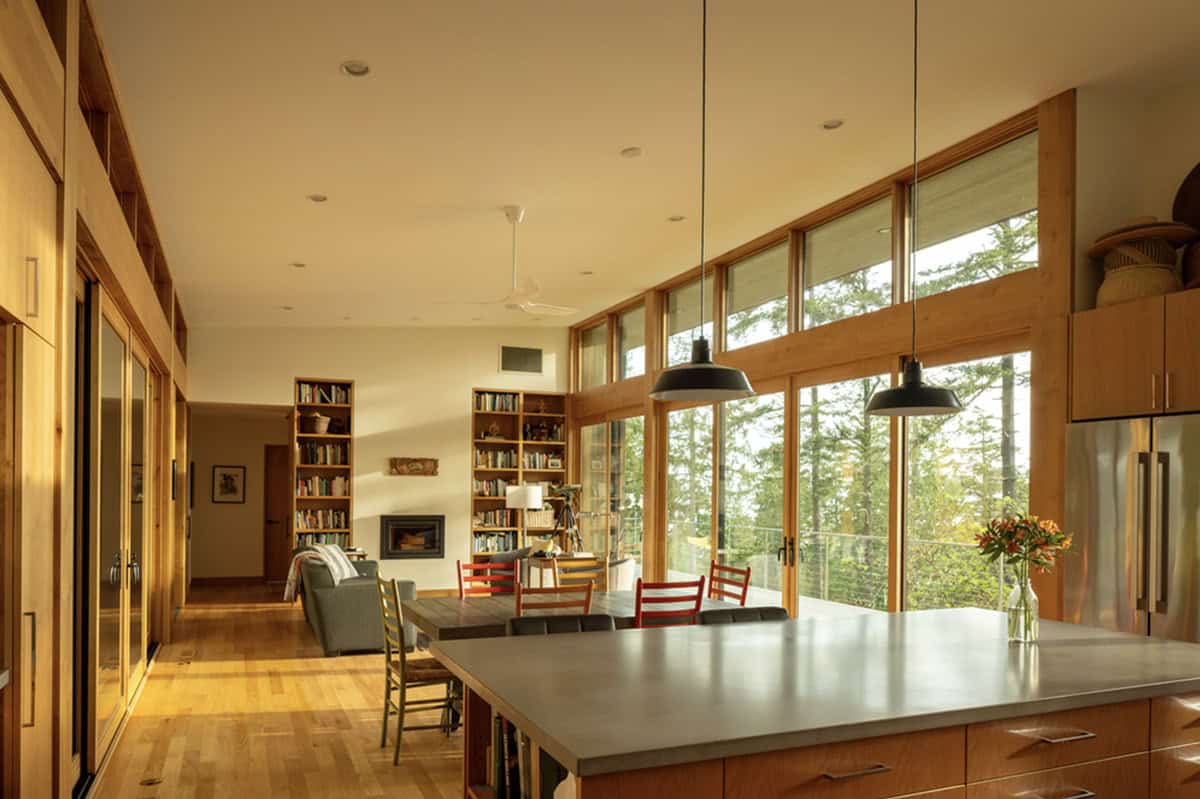 Open-plan kitchen and living area with wood cabinetry, pendant lights, and clerestory windows facing forest