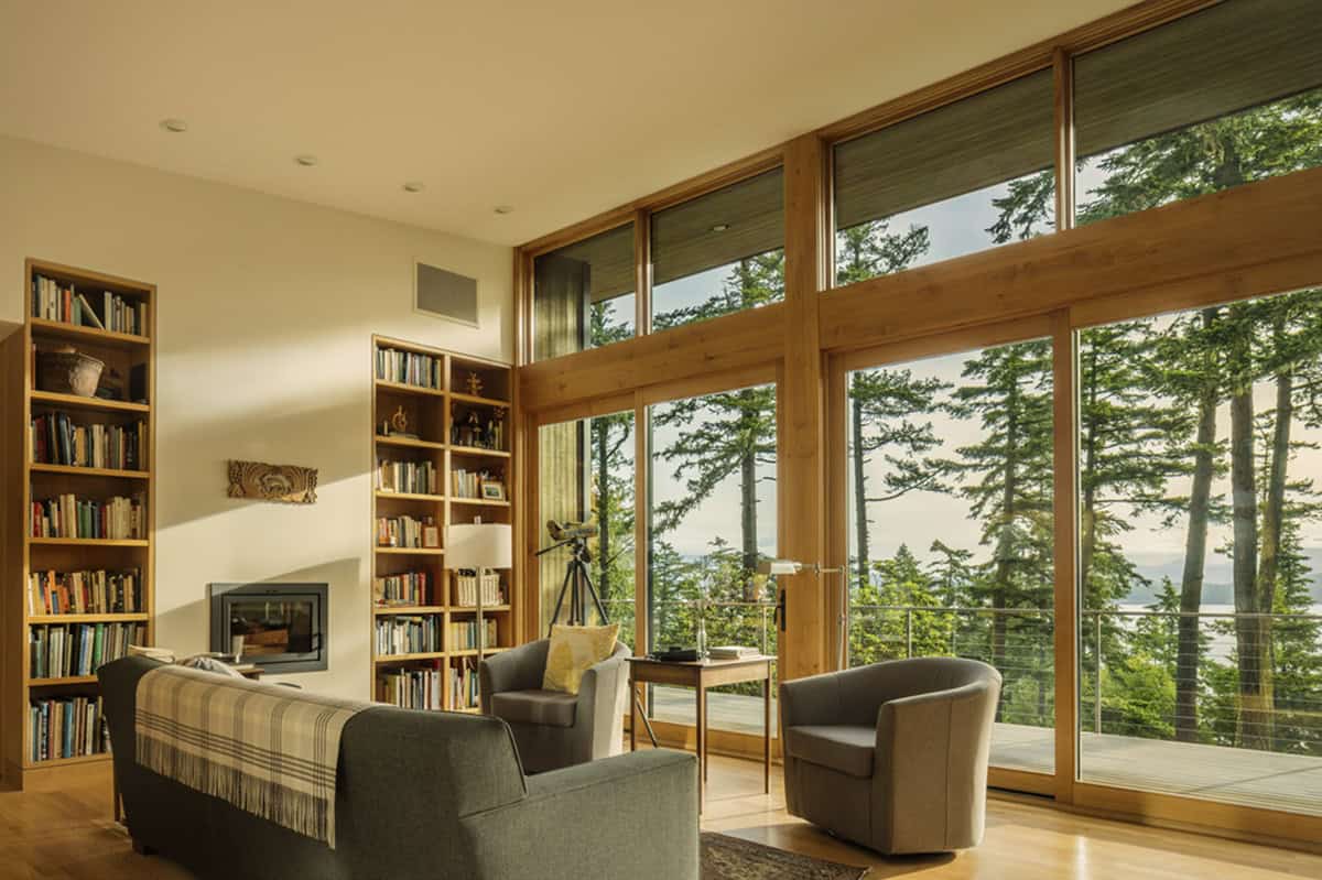 Living room with floor-to-ceiling windows, built-in bookshelves, fireplace, and distant water views through trees