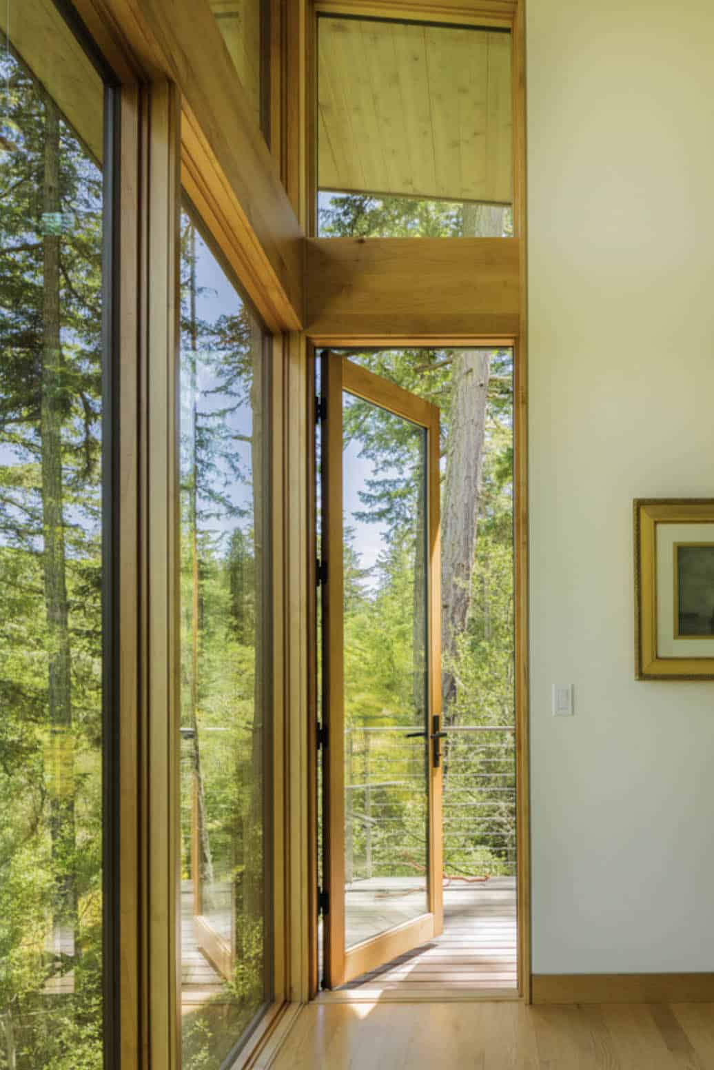 Corner floor-to-ceiling windows with open wood-framed door leading to cable-railed deck among fir trees
