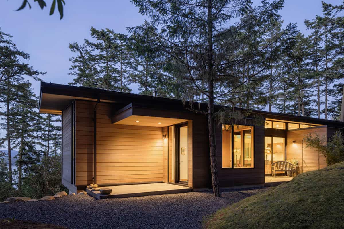Twilight view of home entry with warm interior glow, cedar cladding, and water glimpsed through trees
