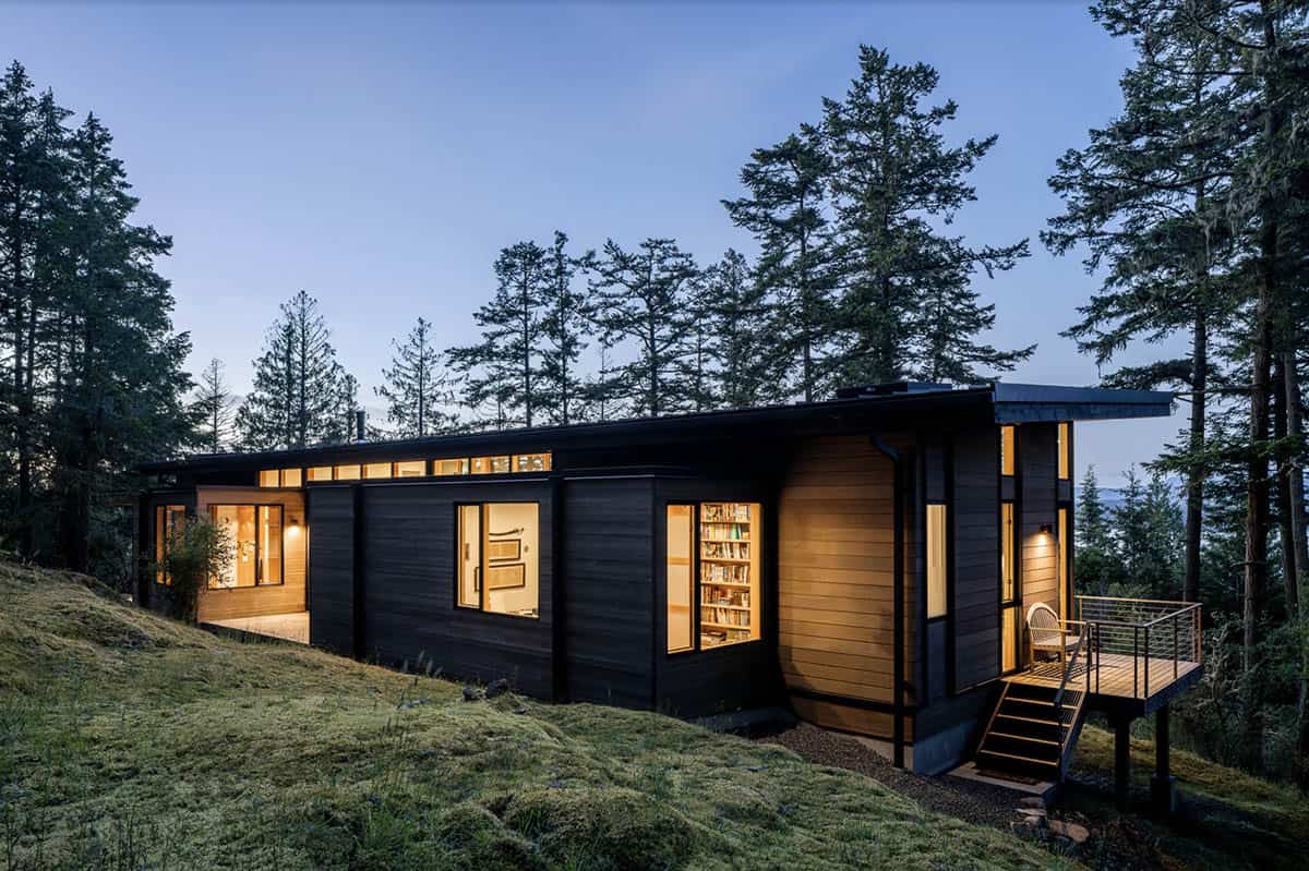 Evening exterior with charred-wood siding, clerestory strip windows, lit bookshelves, and deck with stairs