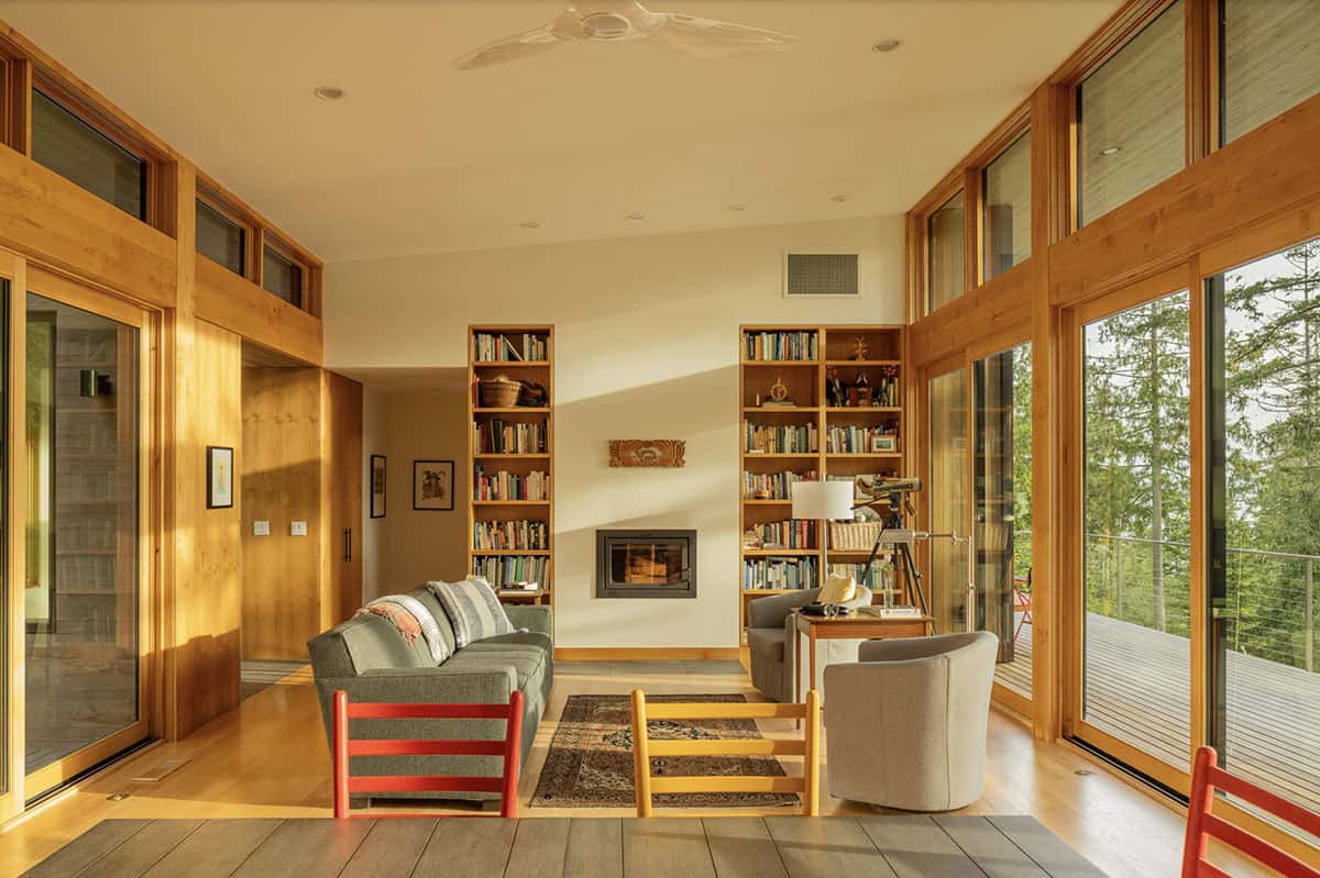 Sun-filled living room with built-in bookshelves, fireplace, colorful chairs, and floor-to-ceiling windows on two sides