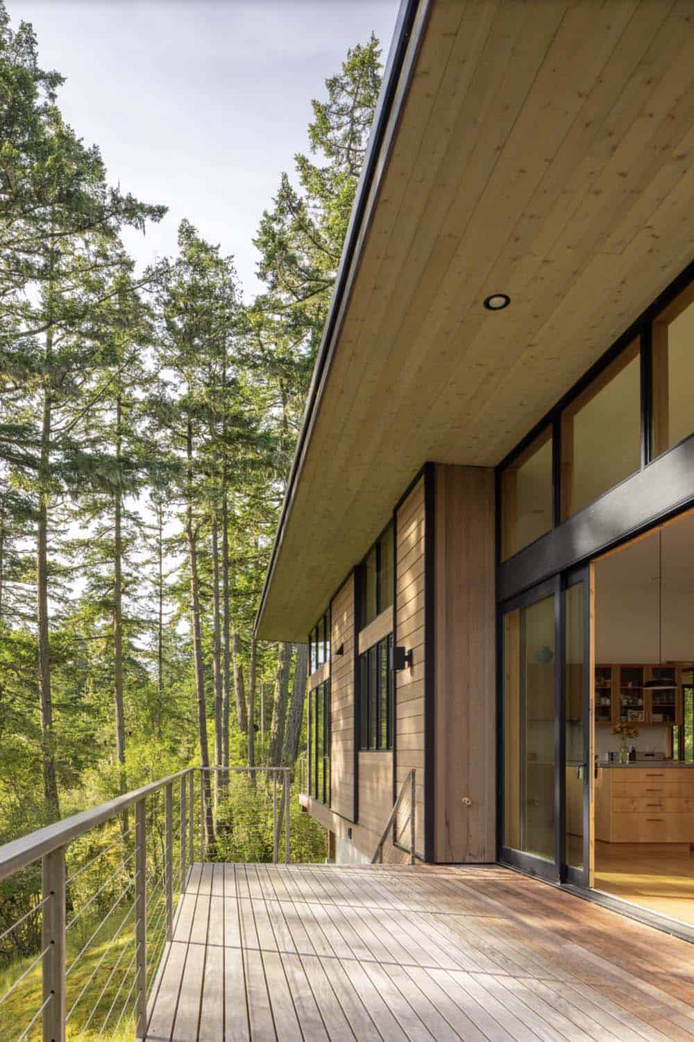 Long wood deck with cable railing under broad roof overhang, sliding glass door open to kitchen beyond