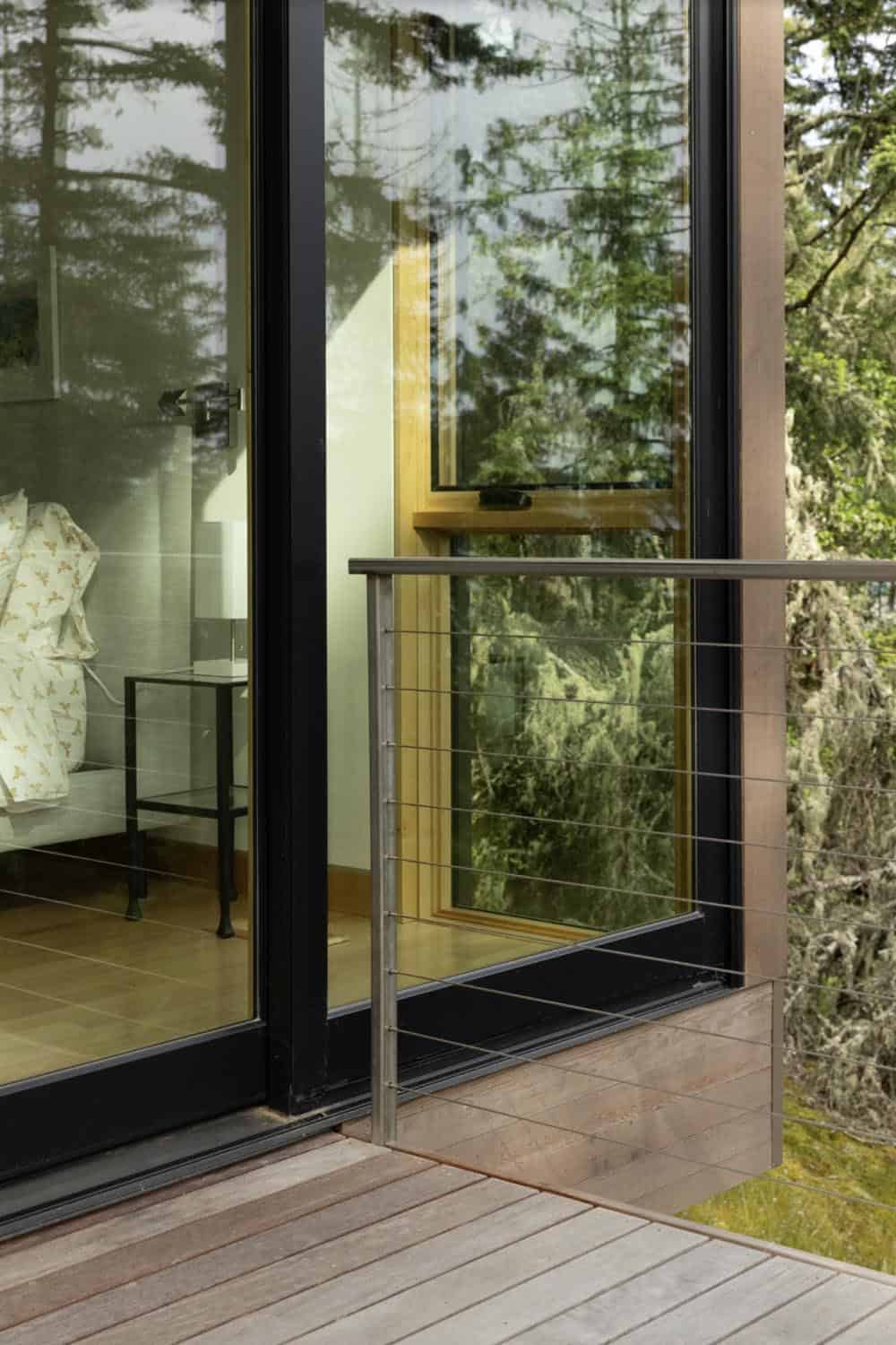 Exterior deck detail with cable railing and black-framed sliding glass door reflecting bedroom interior and forest