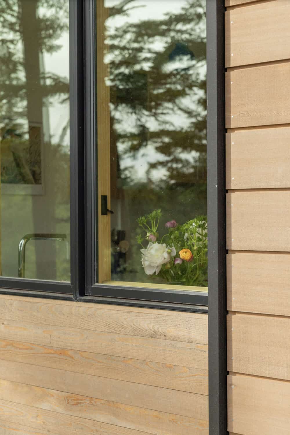 Fresh-cut white peony and ranunculus in vase on windowsill of black-framed window with cedar siding detail