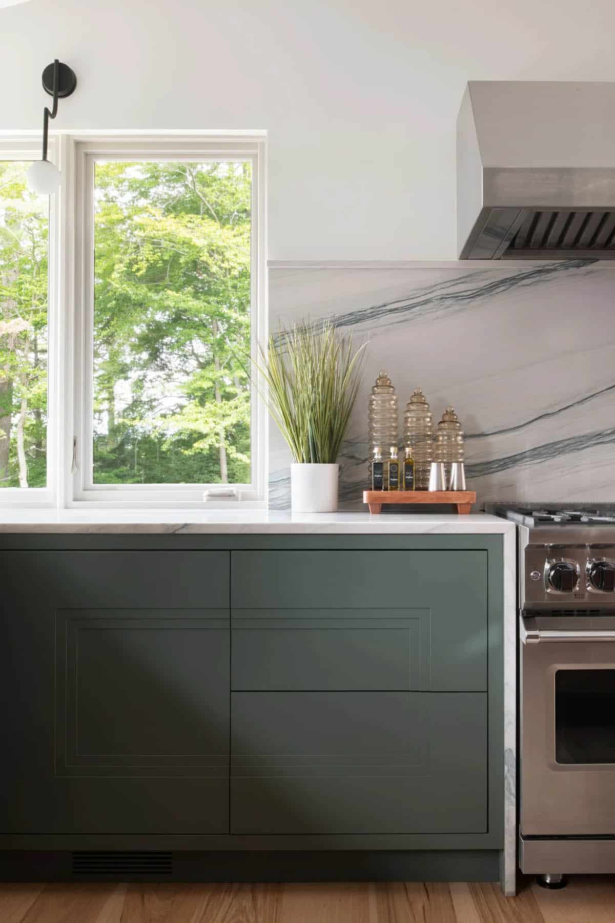 Close-up of dark green kitchen cabinets with marble countertop, professional range, and garden window view