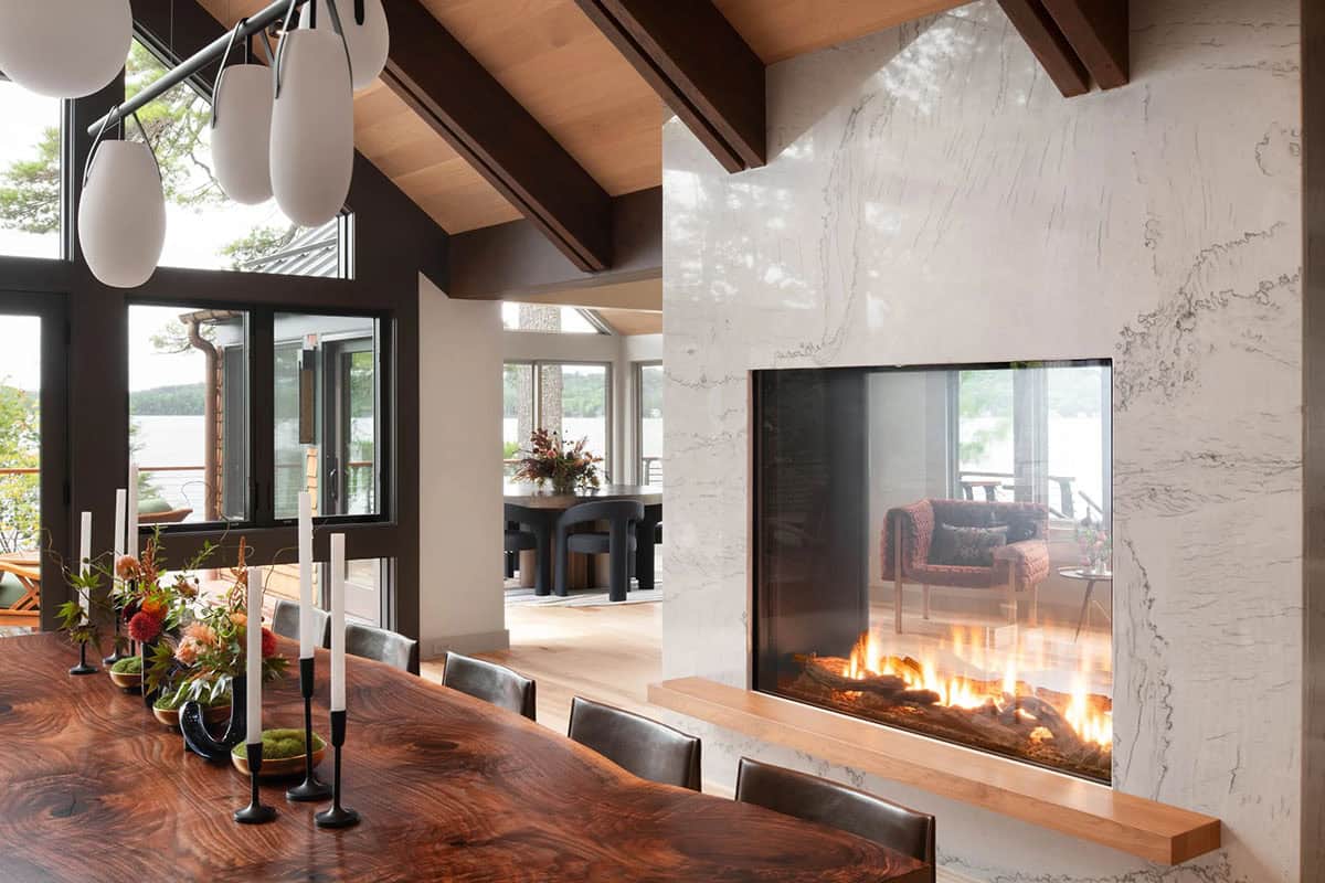 See-through marble fireplace between dining and living areas with lake views and dark wood beams