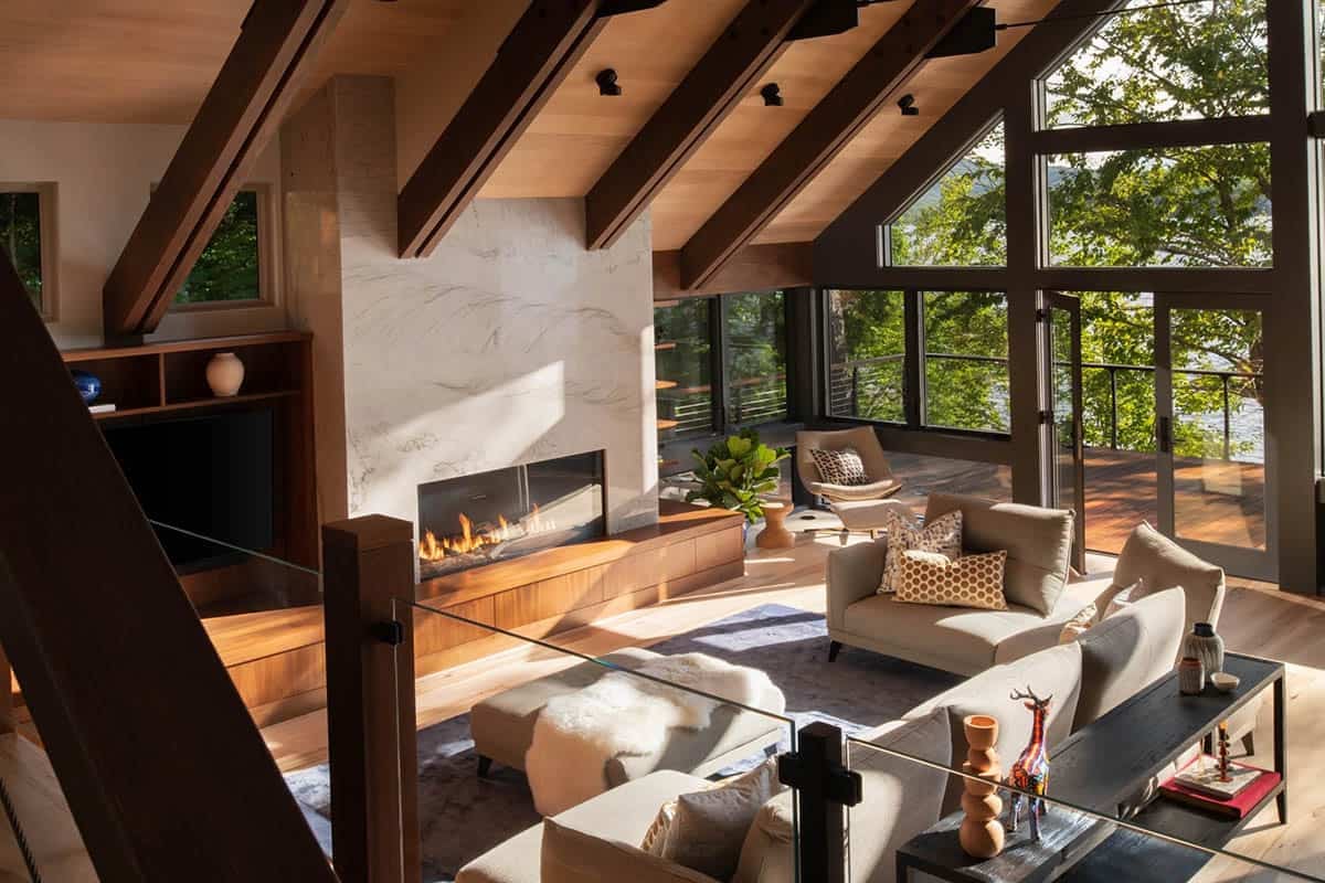Living room with vaulted ceiling, marble fireplace, gray sofas, and panoramic lake views through glass walls