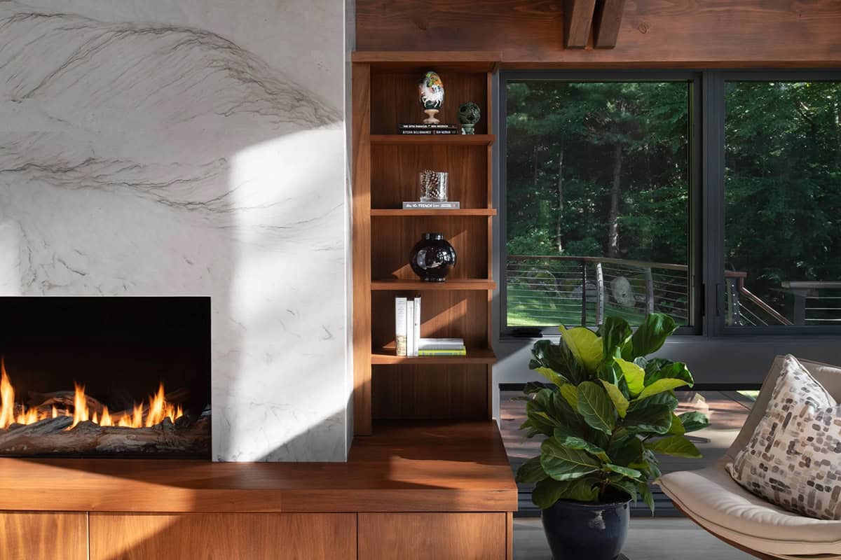 Detail of marble fireplace surround, walnut built-in shelving, and large window with forest view