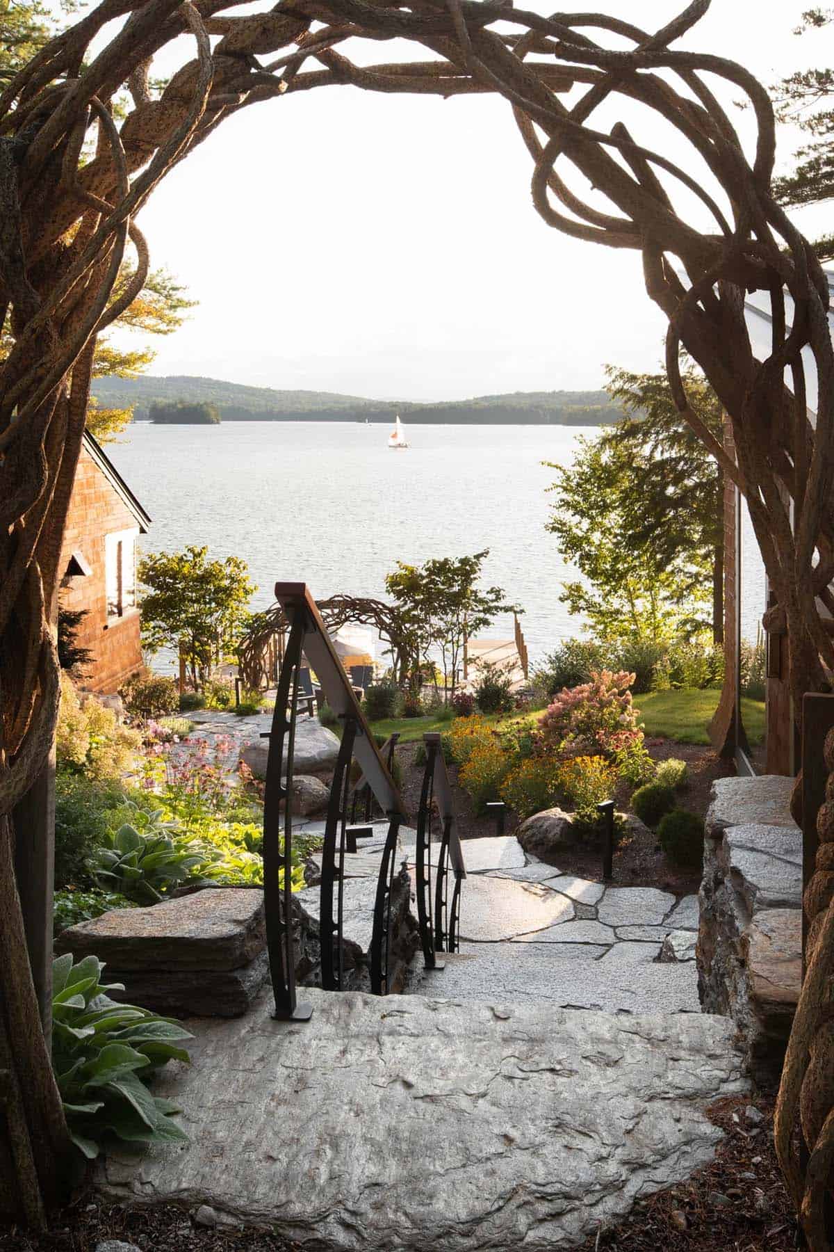 Stone pathway framed by gnarled branch archway leading down to lake with sailboat and lush gardens