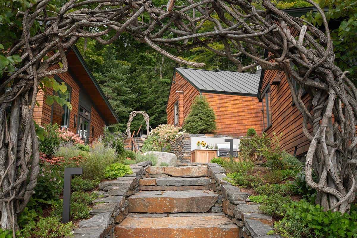 Cedar-clad lakehouse exterior viewed through rustic branch archway with stone steps and landscaped gardens