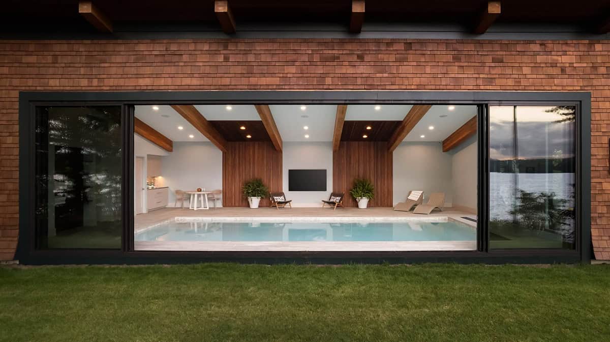 Indoor pool viewed through large picture window at dusk, with wood slat accent wall and TV