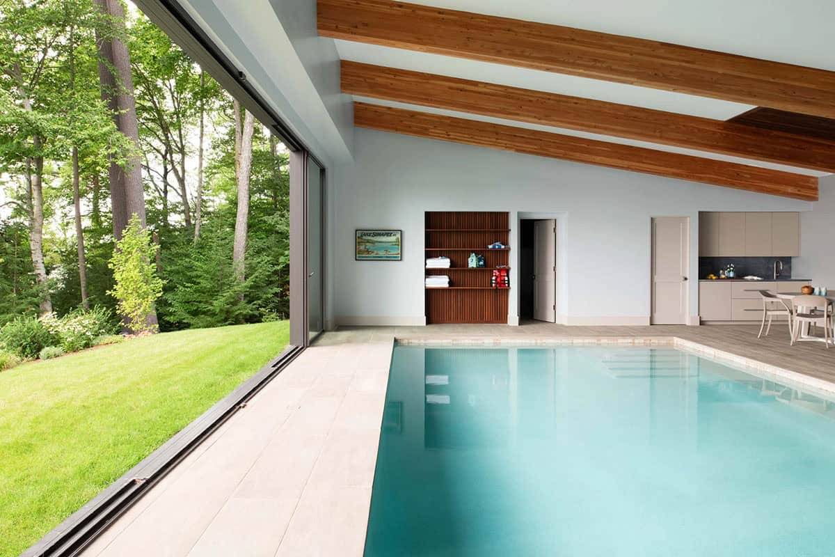 Indoor lap pool with wood ceiling beams, open sliding glass wall to garden, and poolside kitchenette