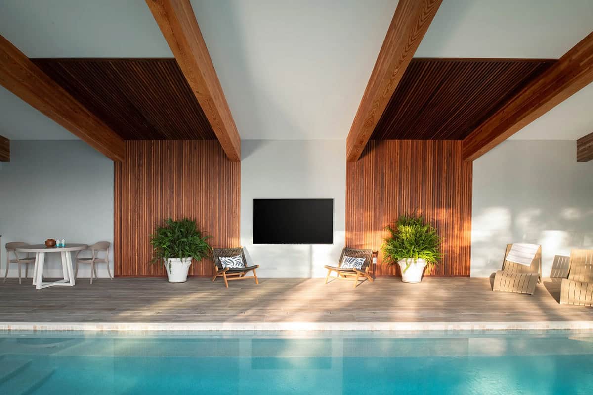 Indoor pool deck with wood slat feature wall, mounted TV, lounge chairs, and ferns in white planters