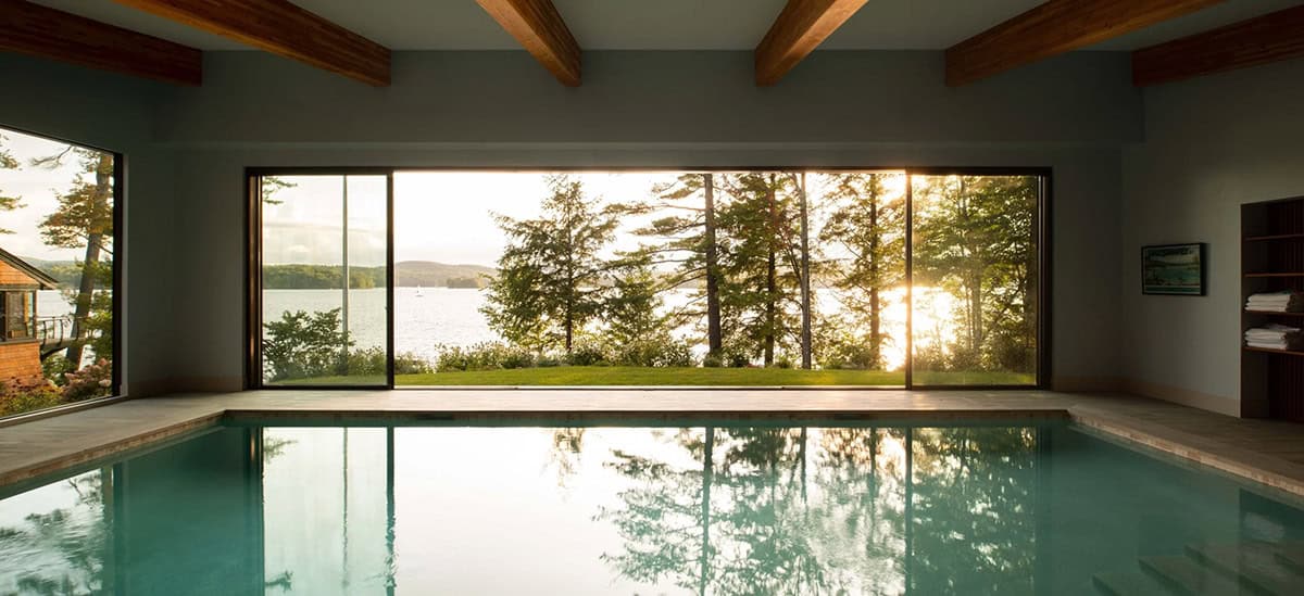 Indoor pool at sunset with panoramic glass wall framing lake and pine trees
