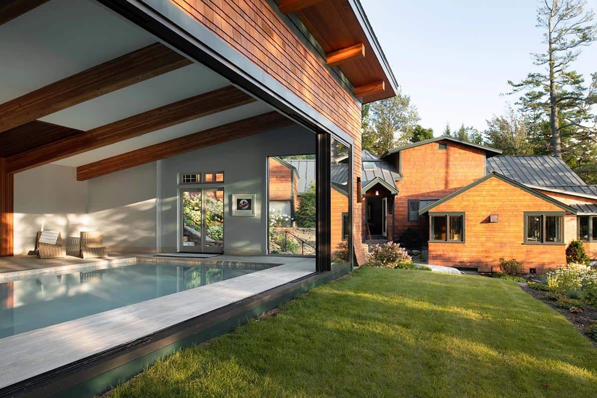Pool house with open sliding wall revealing indoor pool, with cedar-shingled main house beyond