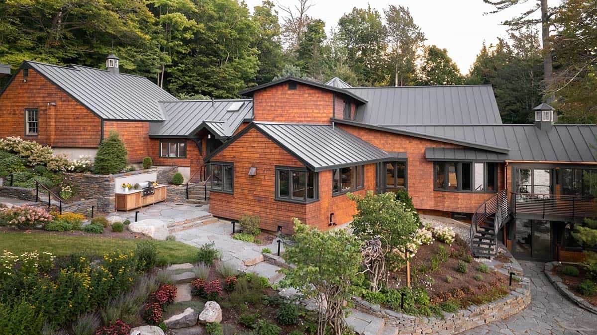 Aerial view of cedar-shingled lakehouse compound with metal roofs, stone terraces, and outdoor kitchen