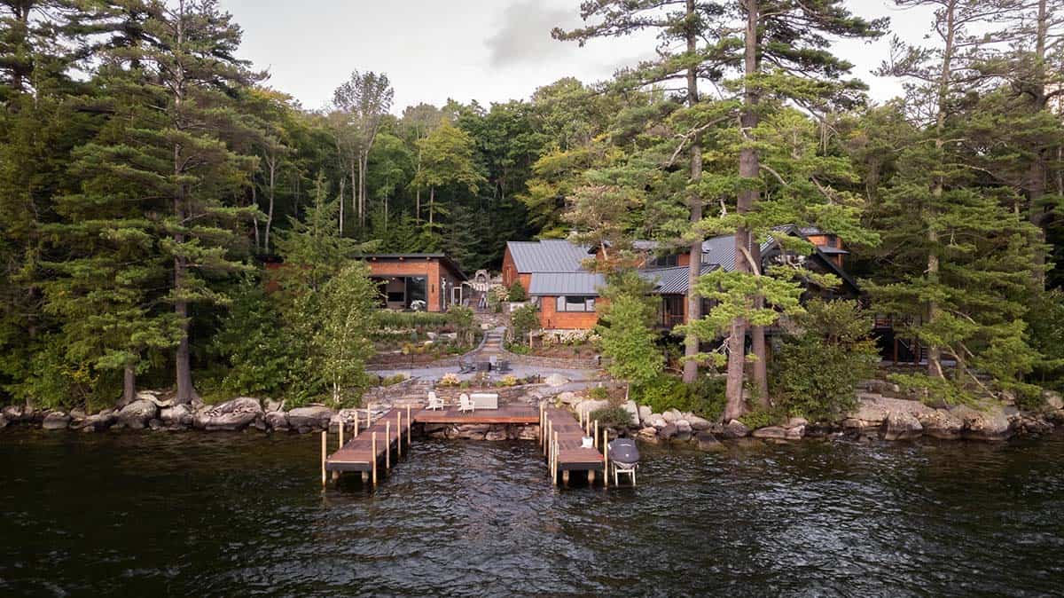 Aerial view from lake showing cedar-shingled compound nestled among pine trees with wood dock