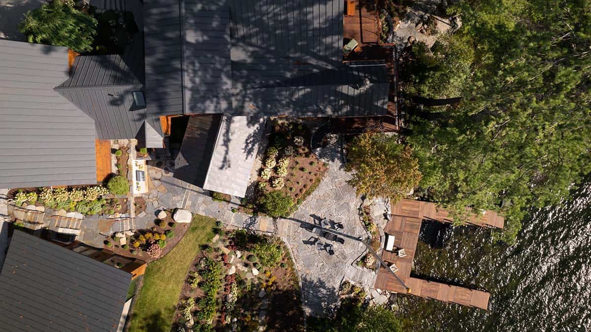 Overhead drone view of lakehouse rooftops, stone patio with fire pit, dock, and shoreline gardens