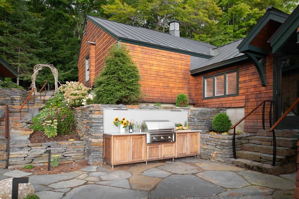 Outdoor kitchen with teak cabinetry and stainless grill on stone patio, cedar-shingled house beyond