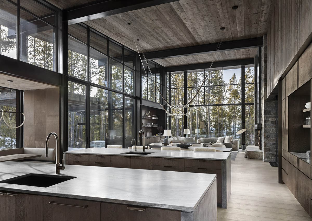 Open-plan kitchen and living area with double islands, steel-framed glass walls, and lodgepole pine forest views