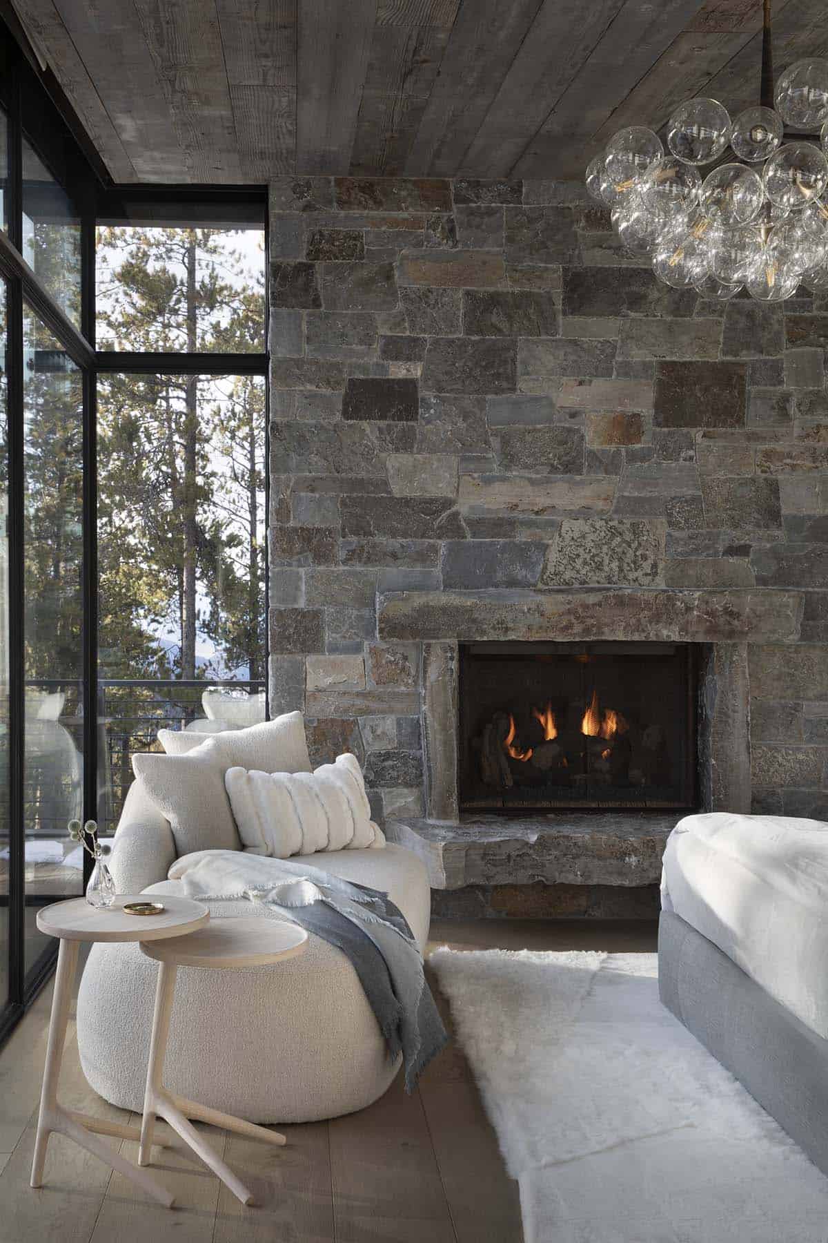 Primary bedroom sitting area with bouclé chair, lit stone fireplace, bubble glass chandelier, and forest views