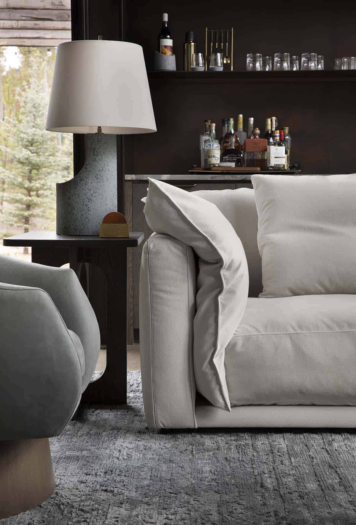 Living room detail of cream sofa, speckled ceramic table lamp, and dark shelved bar with glassware