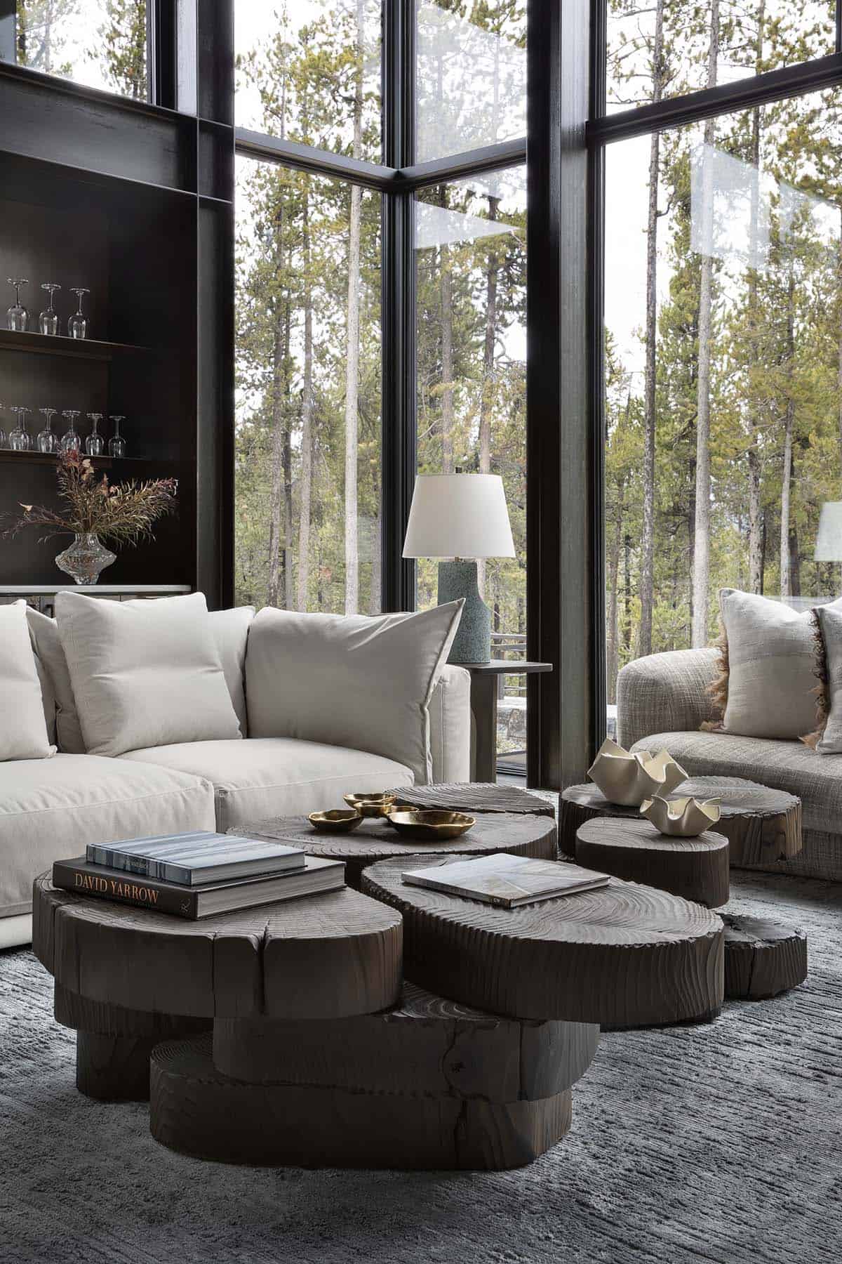 Living room with cream sofa, cluster of dark wood stump coffee tables, and floor-to-ceiling forest views