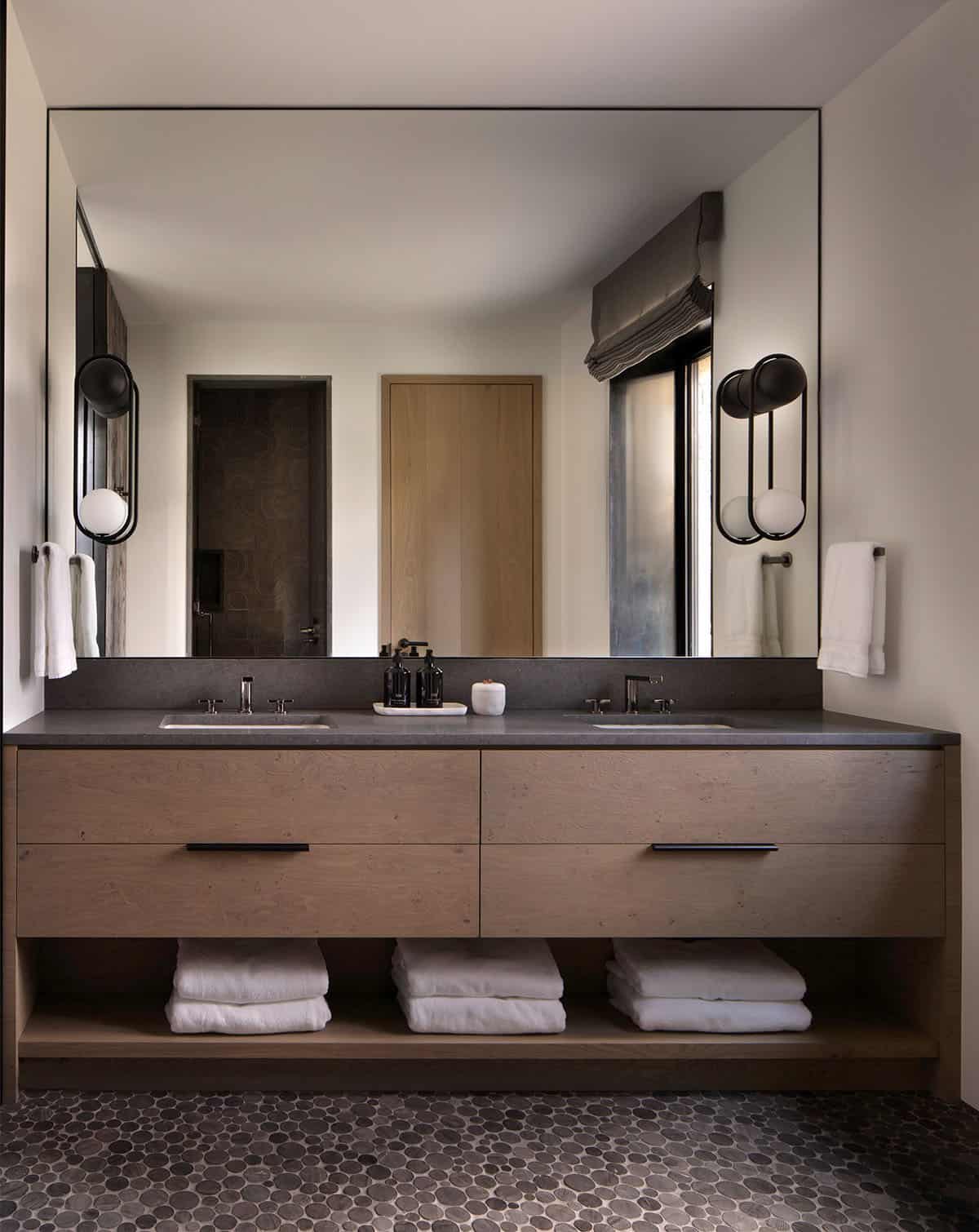 Guest bathroom with double undermount sinks, dark stone countertop, large mirror, and pebble tile floor