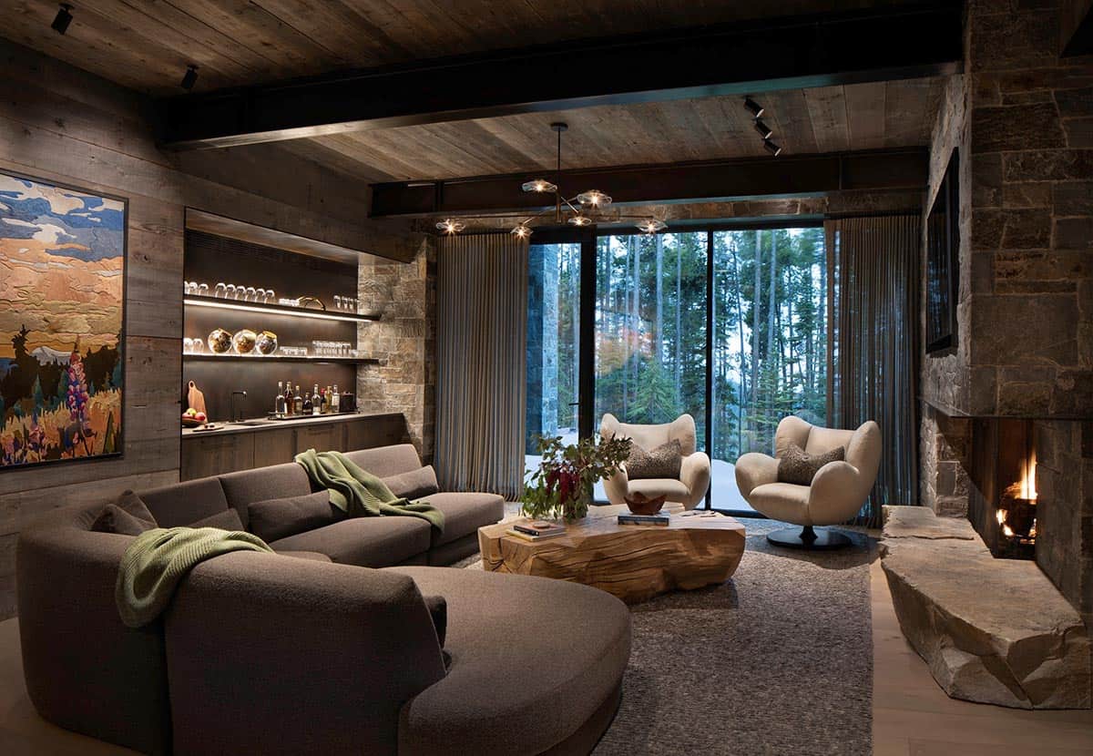 Lower-level bar and lounge with curved sectional, stone fireplace, lit bar shelving, and forest views at night