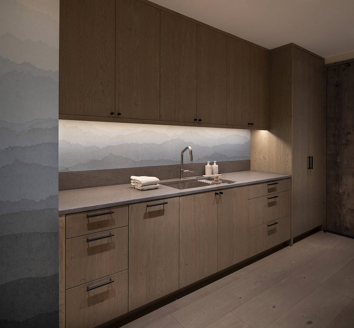 Laundry room with warm oak cabinetry, under-cabinet lighting, mountain mural backsplash, and stone countertop