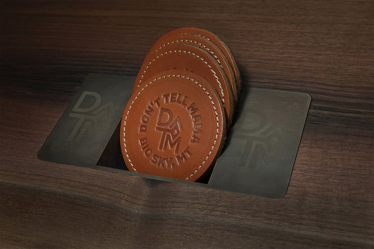 Close-up of stamped leather coasters reading "Don't Tell Mama, Big Sky MT" on dark wood bar surface