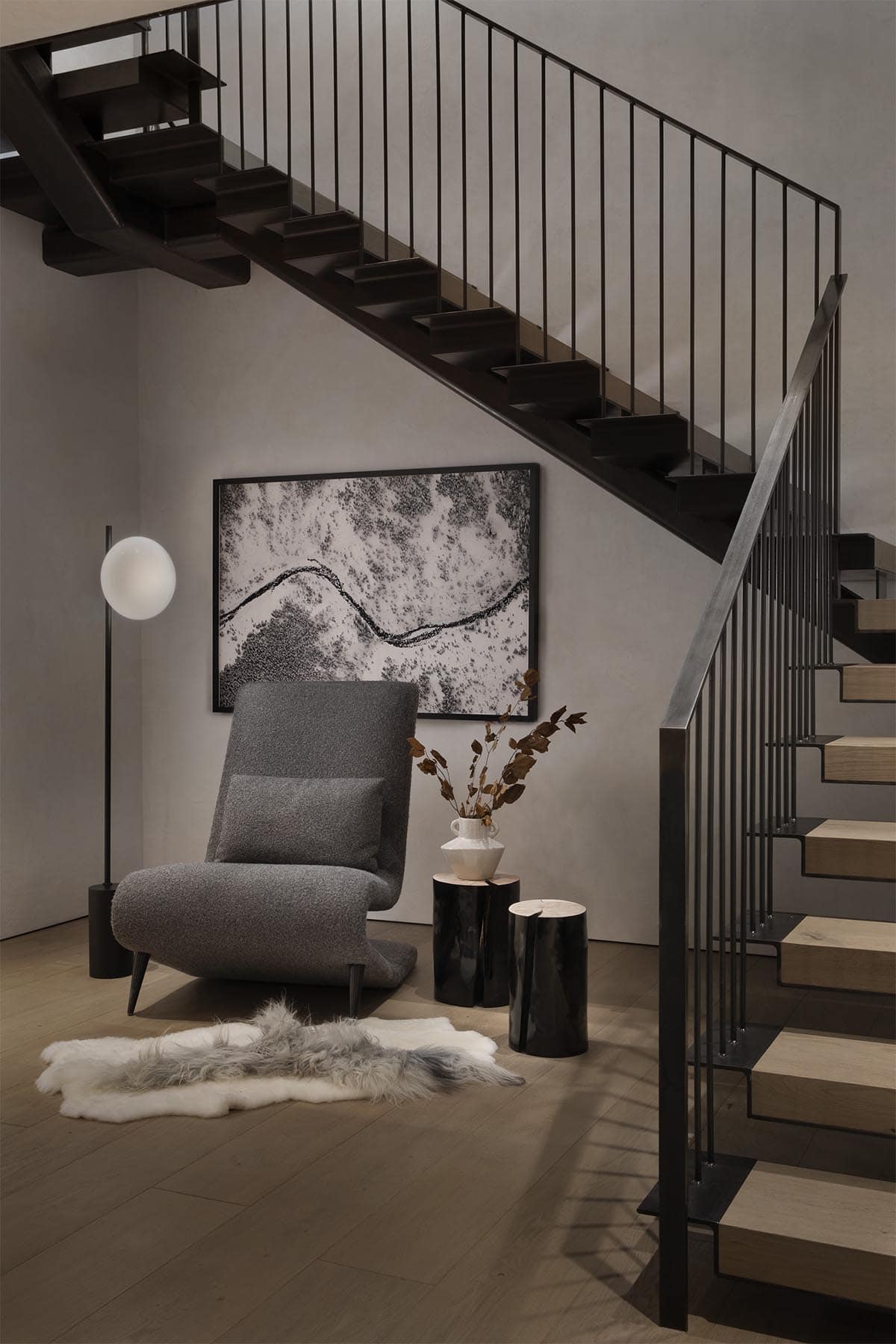 Floating steel staircase landing with bouclé lounge chair, globe floor lamp, log side tables, and aerial landscape art