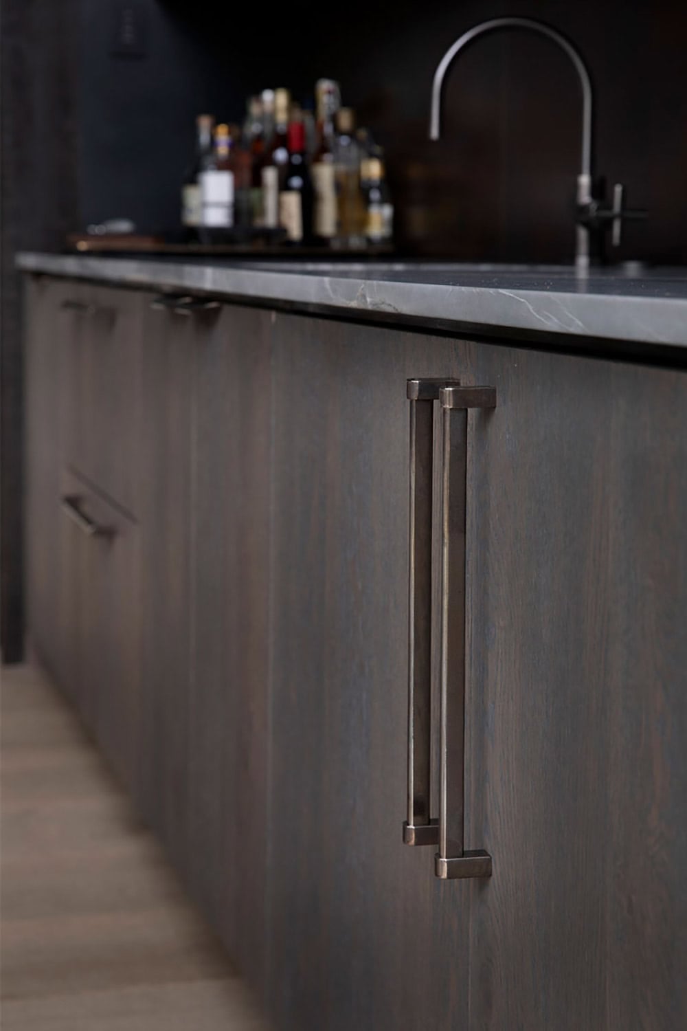 Bar cabinet detail with dark oak doors, aged brass hardware pulls, dark stone countertop, and liquor bottles beyond