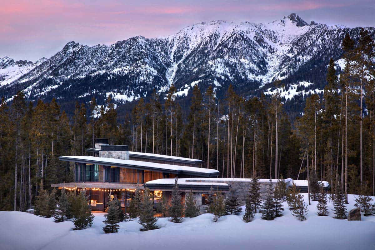 Twilight exterior of modern mountain home with snow-covered peaks, string lights, and warm interior glow