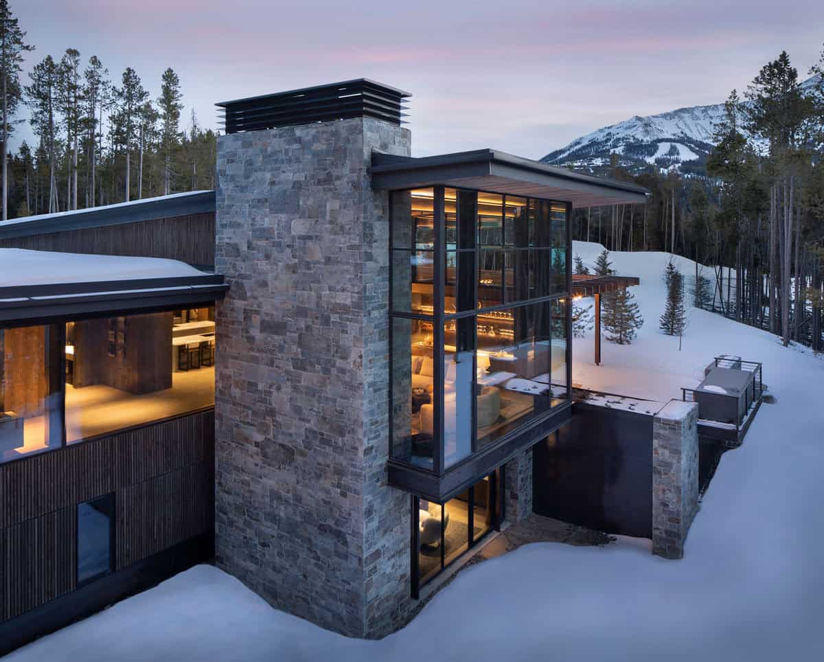 Dusk exterior detail of stone chimney tower with illuminated double-height steel-framed windows and snowy surroundings