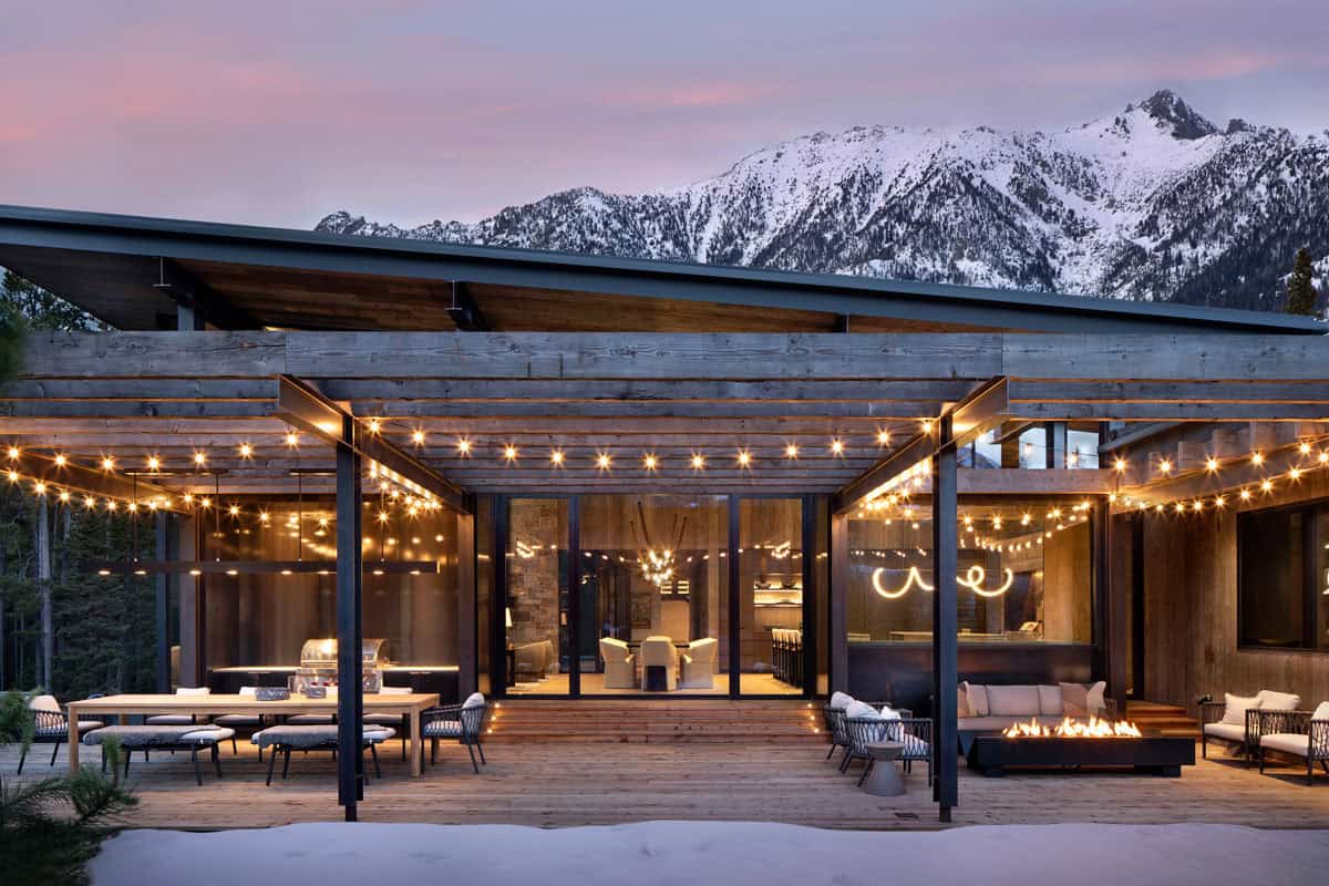 Covered outdoor entertaining deck with string lights, fire pit, dining table, and snowy mountain backdrop at dusk