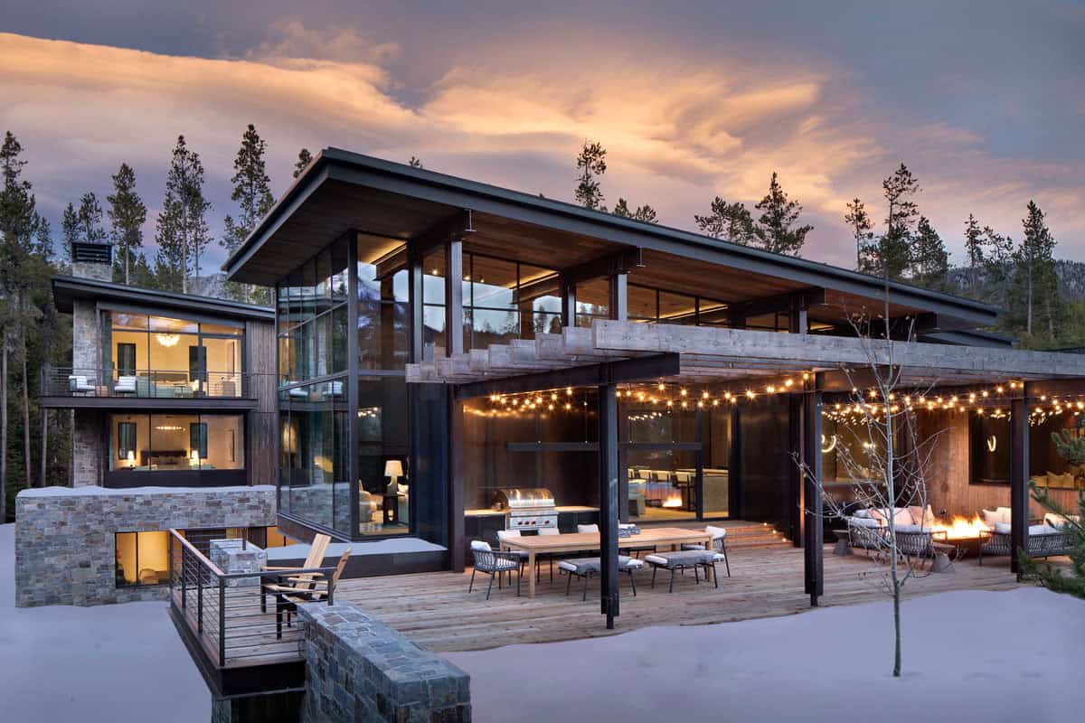 Full rear exterior at dusk showing stone and wood facade, lit outdoor deck, fire pit, and glowing interiors