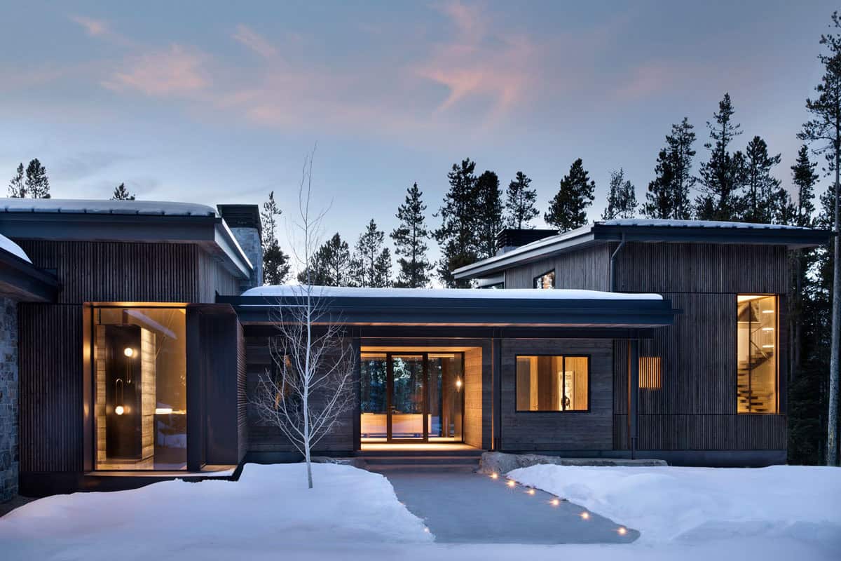 Winter twilight front entry with vertical slatted wood siding, lit pathway, bare aspen tree, and warm interior glow