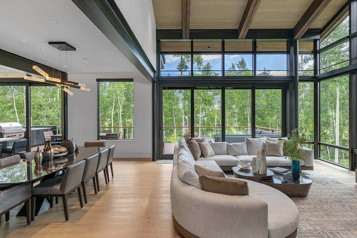 Open-plan living and dining area with curved sectional sofa and double-height windows overlooking aspen forest