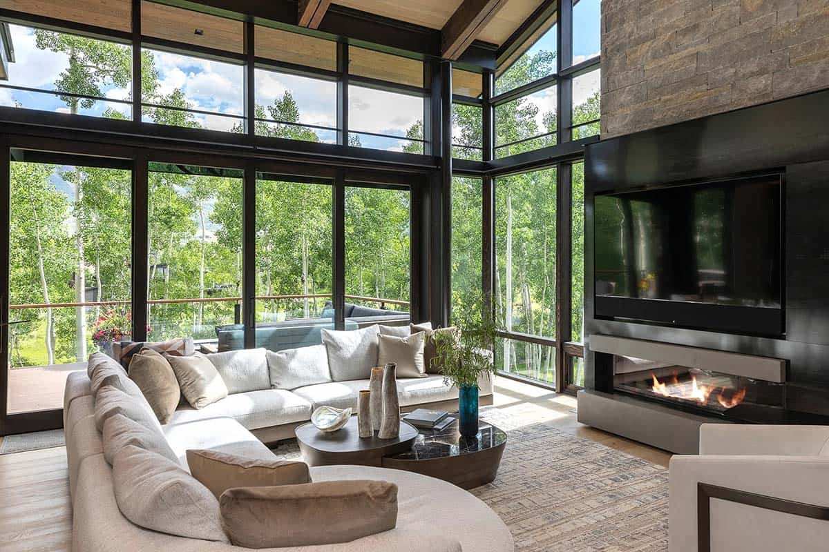 Living room corner with curved sectional, stone fireplace, and two-story glass walls framing aspen trees