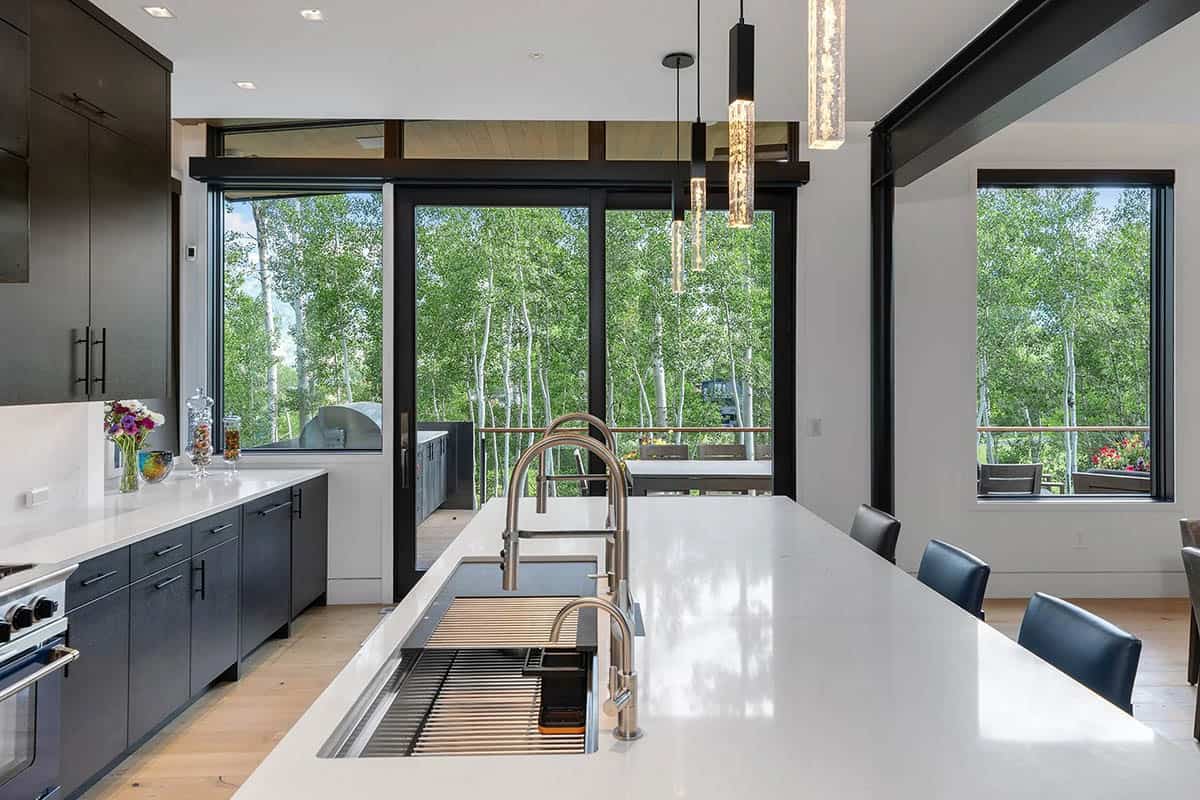 Modern kitchen island with white quartz countertop, dual faucets, dark cabinets, and forest views