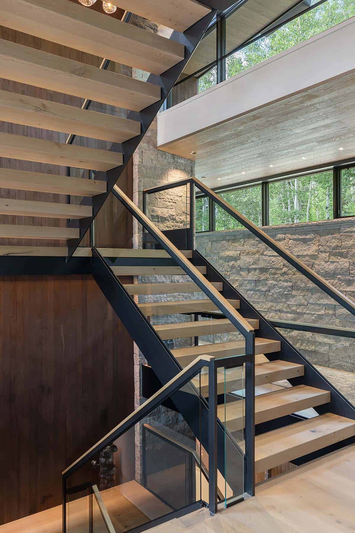 Open-riser staircase with light wood treads, black steel frame, and glass railings against stone wall