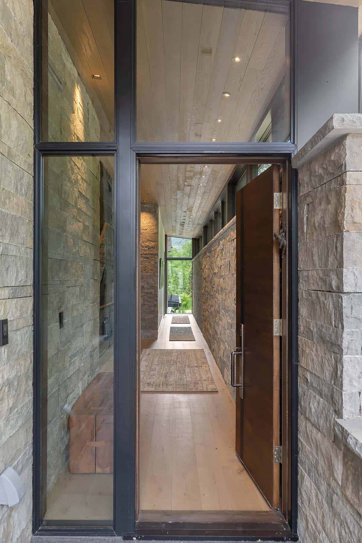 Entry corridor with pivot wood door, black steel-framed glass panels, and stacked stone walls