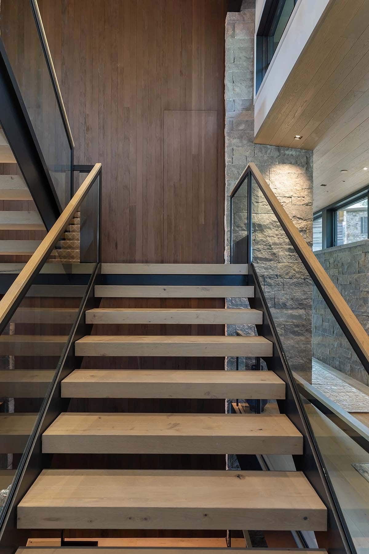 Floating staircase with natural oak treads, black steel stringers, and glass railings beside wood-paneled wall