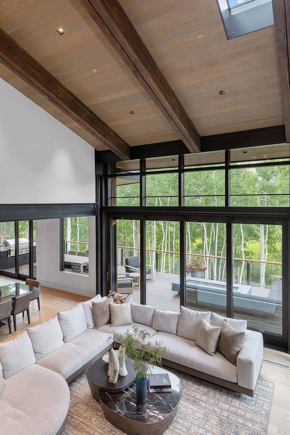 Great room with exposed wood beam ceiling, skylights, sectional sofa, and full-height aspen forest views