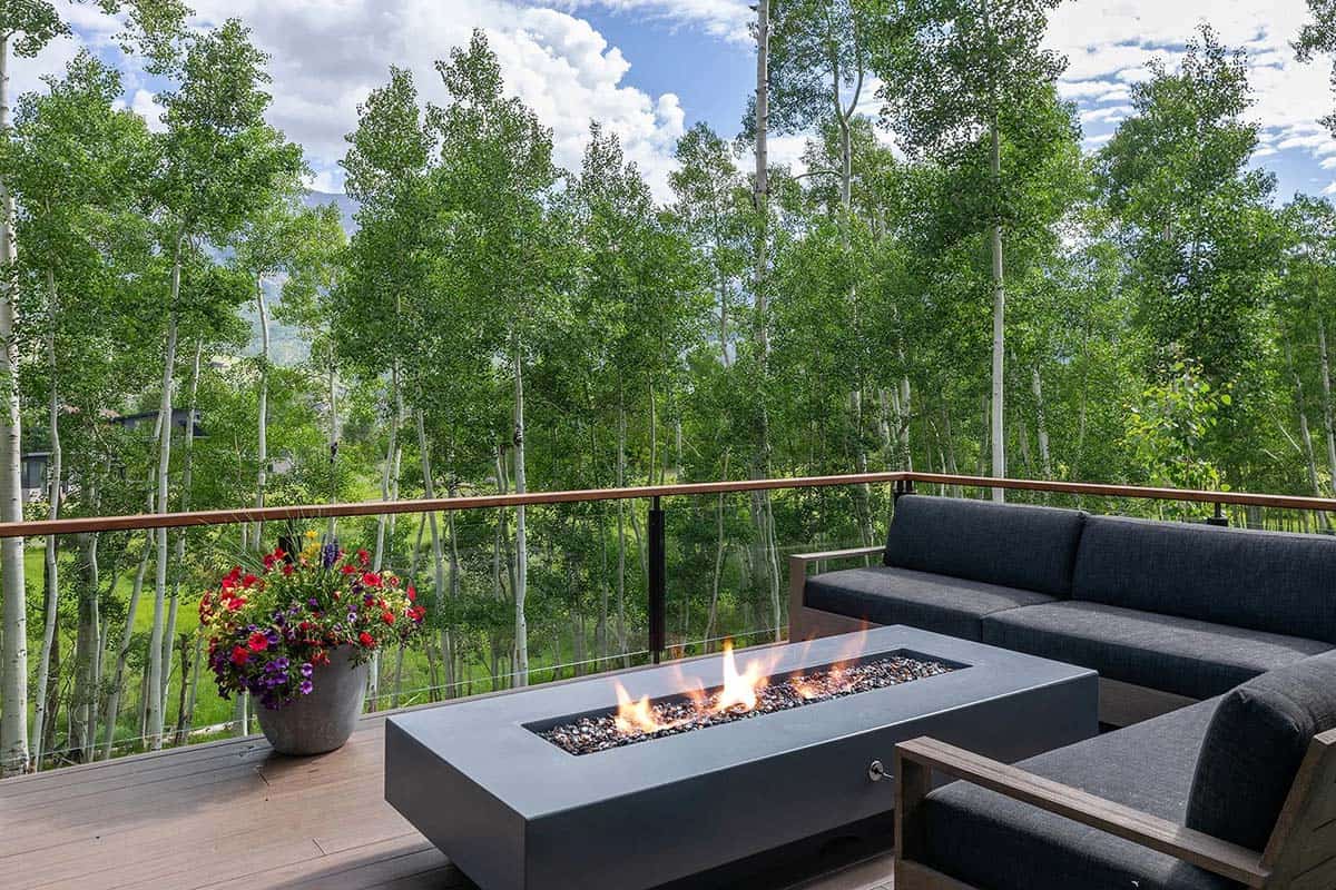 Deck with concrete linear fire table, dark sectional seating, glass railing, and aspen grove backdrop