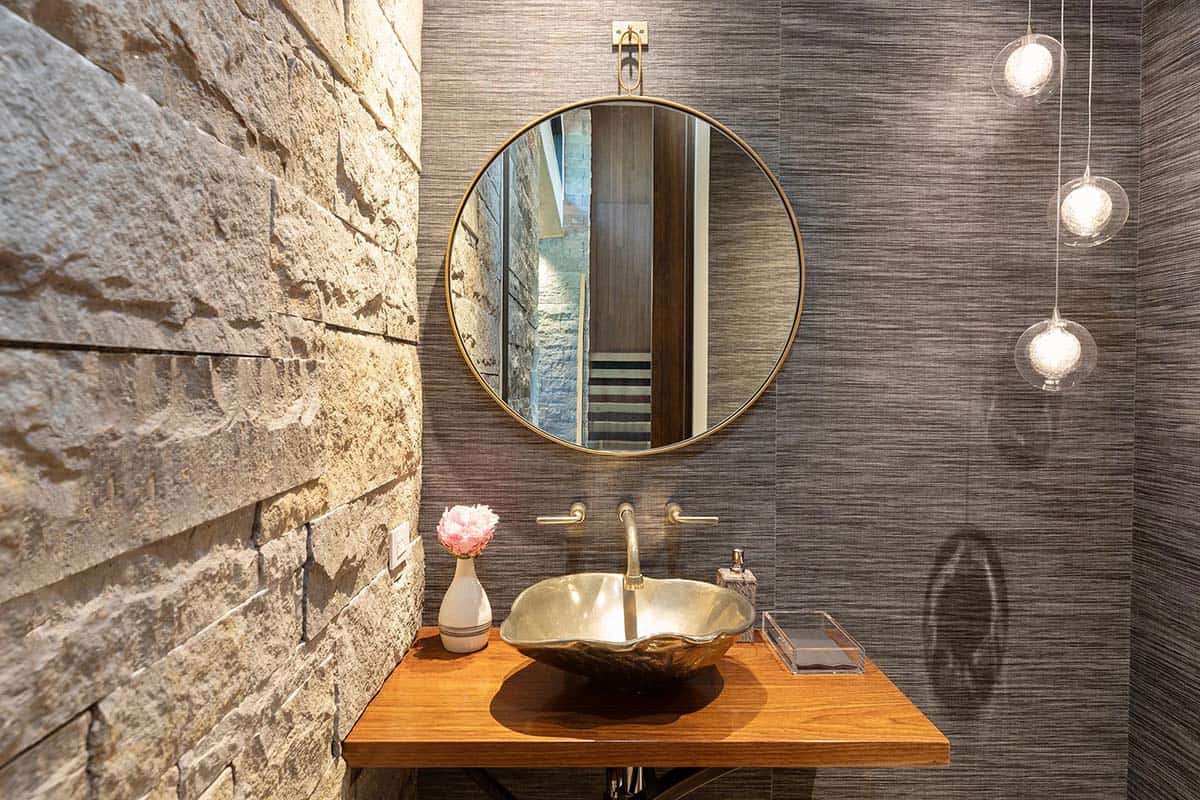 Powder room with hammered bronze vessel sink, round brass-framed mirror, stone wall, and glass pendant lights